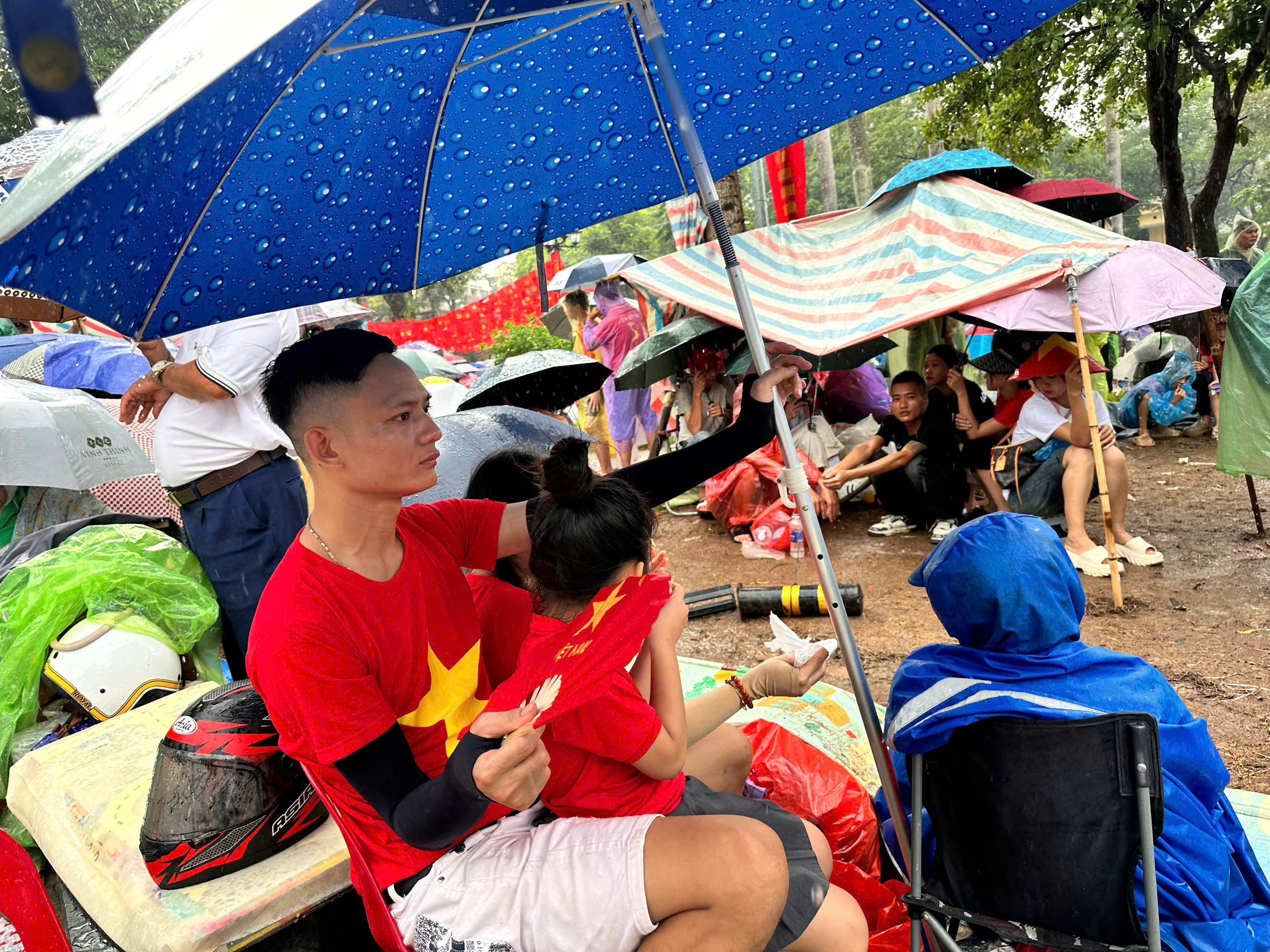 Người bố ôm con gái ngồi trú dưới ô, cùng hòa vào dòng người chờ đợi Người bố ôm con gái ngồi trú dưới ô, cùng hòa vào dòng người chờ đợi